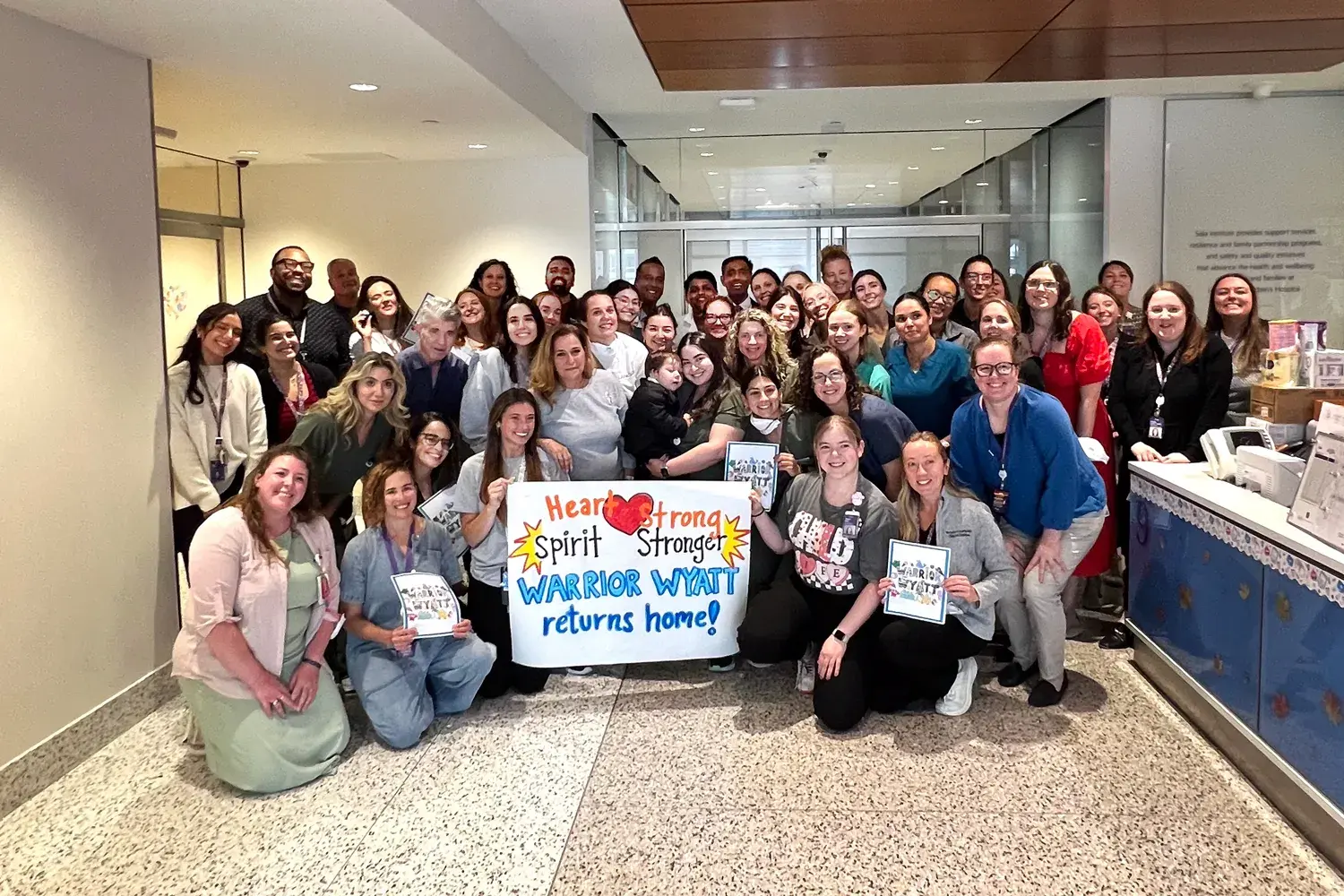 A large group of hospital staff members pose together in a bright hallway, many kneeling in front and others standing behind them. Several people hold handmade signs, including a large central poster that reads “Heart Strong, Spirit Stronger, WARRIOR WYATT returns home!” decorated with colorful drawings and hearts, creating a celebratory send-off scene.
