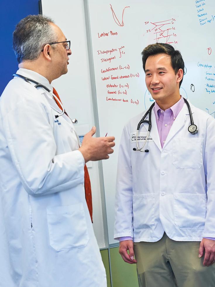 A male NYU Grossman Long Island School of Medicine doctor and male resident wearing white coats talk in a hospital foyer.