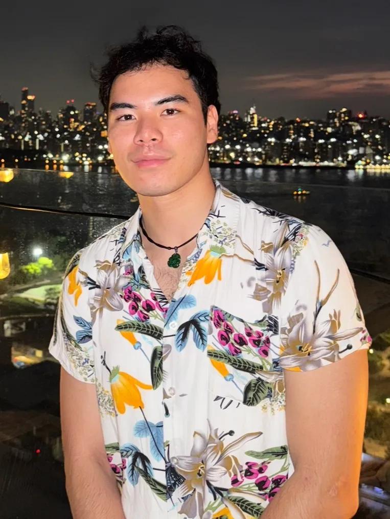 Wei Xie, a student at NYU Grossman School of Medicine, wears a floral button down shirt with an evening view of the New Yory City skyline in the background.