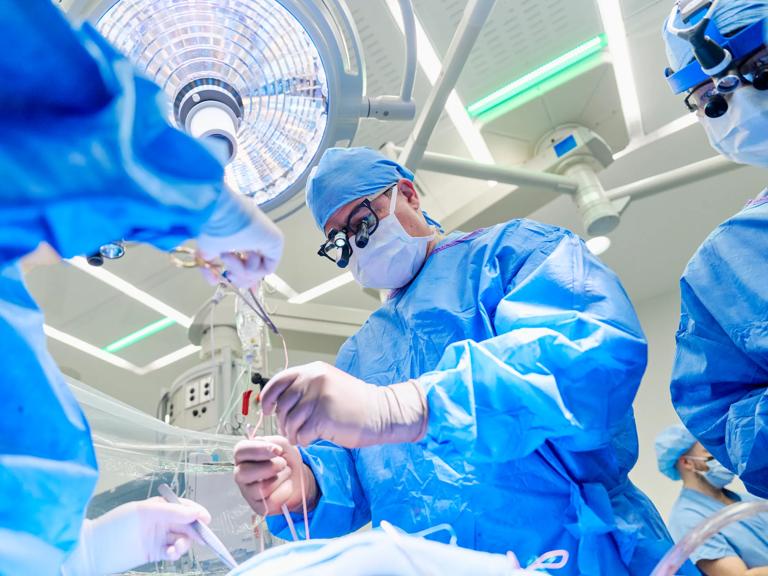 Surgical team in blue sterile gowns and gloves work under bright operating room lights, handling instruments and tubing during a procedure.