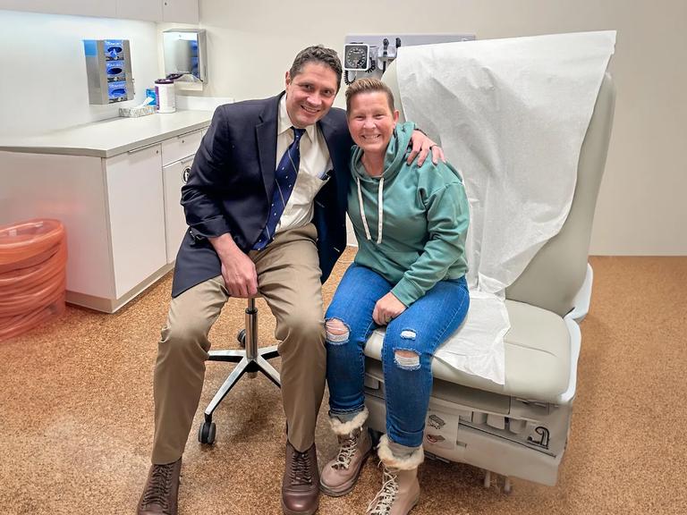 Dr. Wolfgang and Martinez sitting together in exam room with an arm around the other's shoulders and back, both smiling.