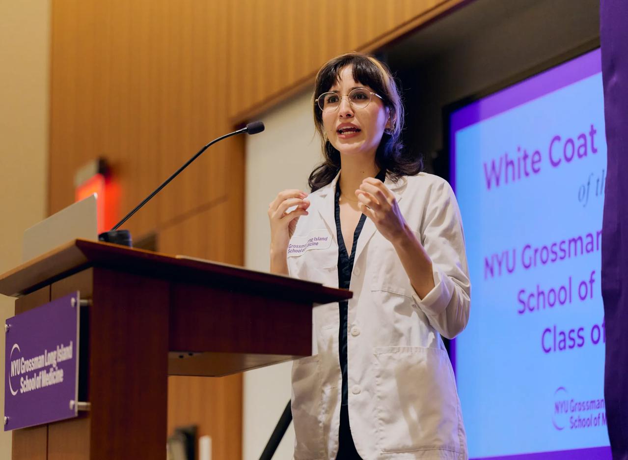 A young female doctor from Grossman Long Island School of Medicine stands and talks at a podium with a photo of a white NYU Langone Health logo on a purple background on the front.