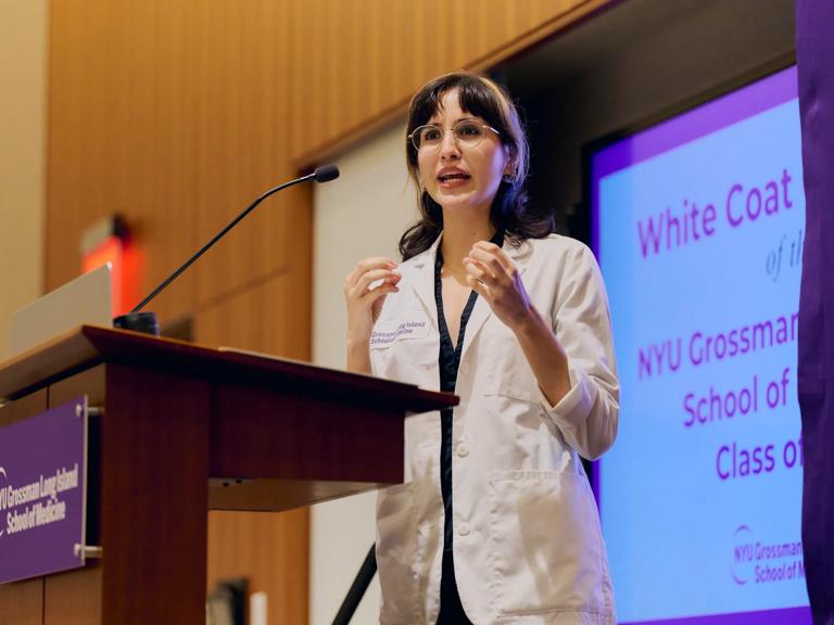 A young female doctor from Grossman Long Island School of Medicine stands and talks at a podium with a photo of a white NYU Langone Health logo on a purple background on the front.