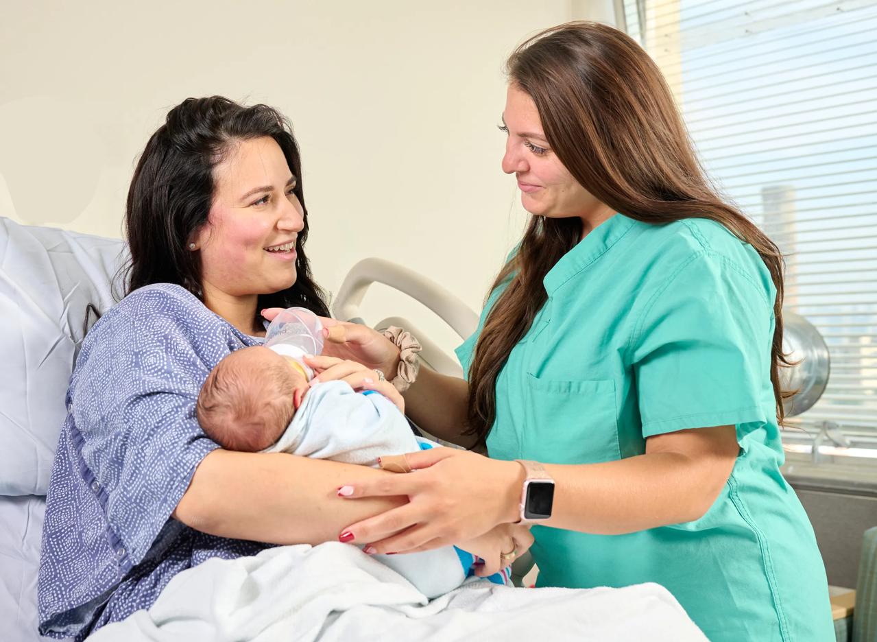 A female patient, left, lies down in a hospital bed while holding a baby. A nurse, right, stands next to the patient and helps feed the baby a bottle.