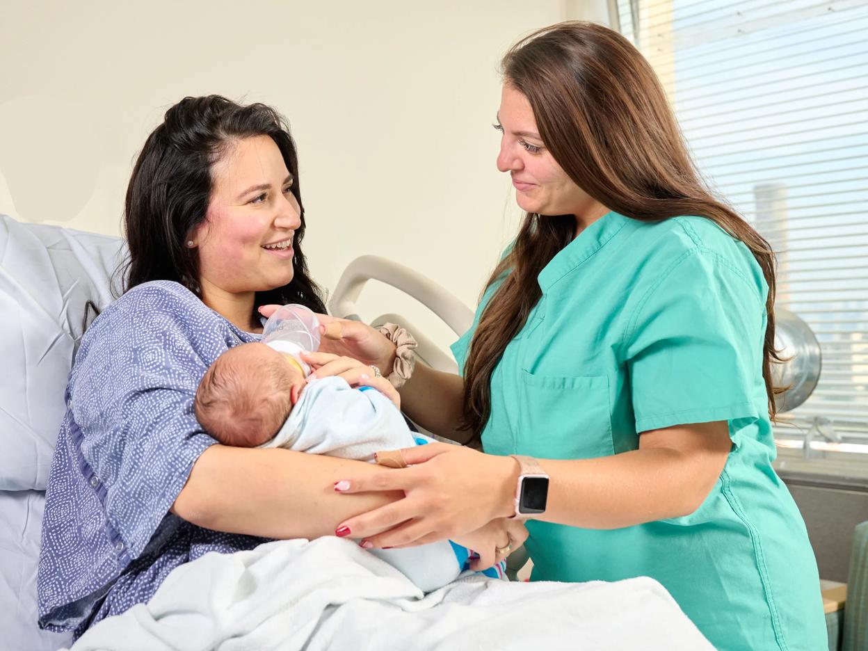 A female patient, left, lies down in a hospital bed while holding a baby. A nurse, right, stands next to the patient and helps feed the baby a bottle.