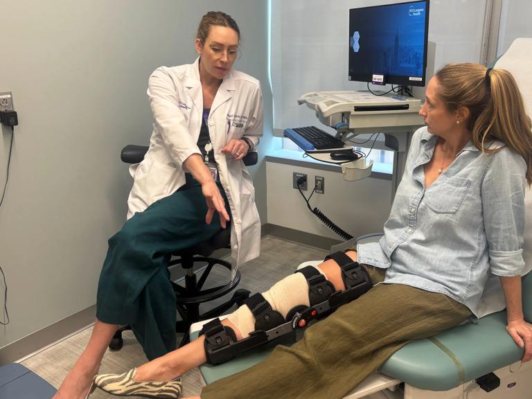 In a medical exam room, a healthcare professional in a white coat sits on a stool and gestures while talking with a patient who is seated on an exam table. Patient has a large black knee brace and bandage on one leg, which is extended onto a support.