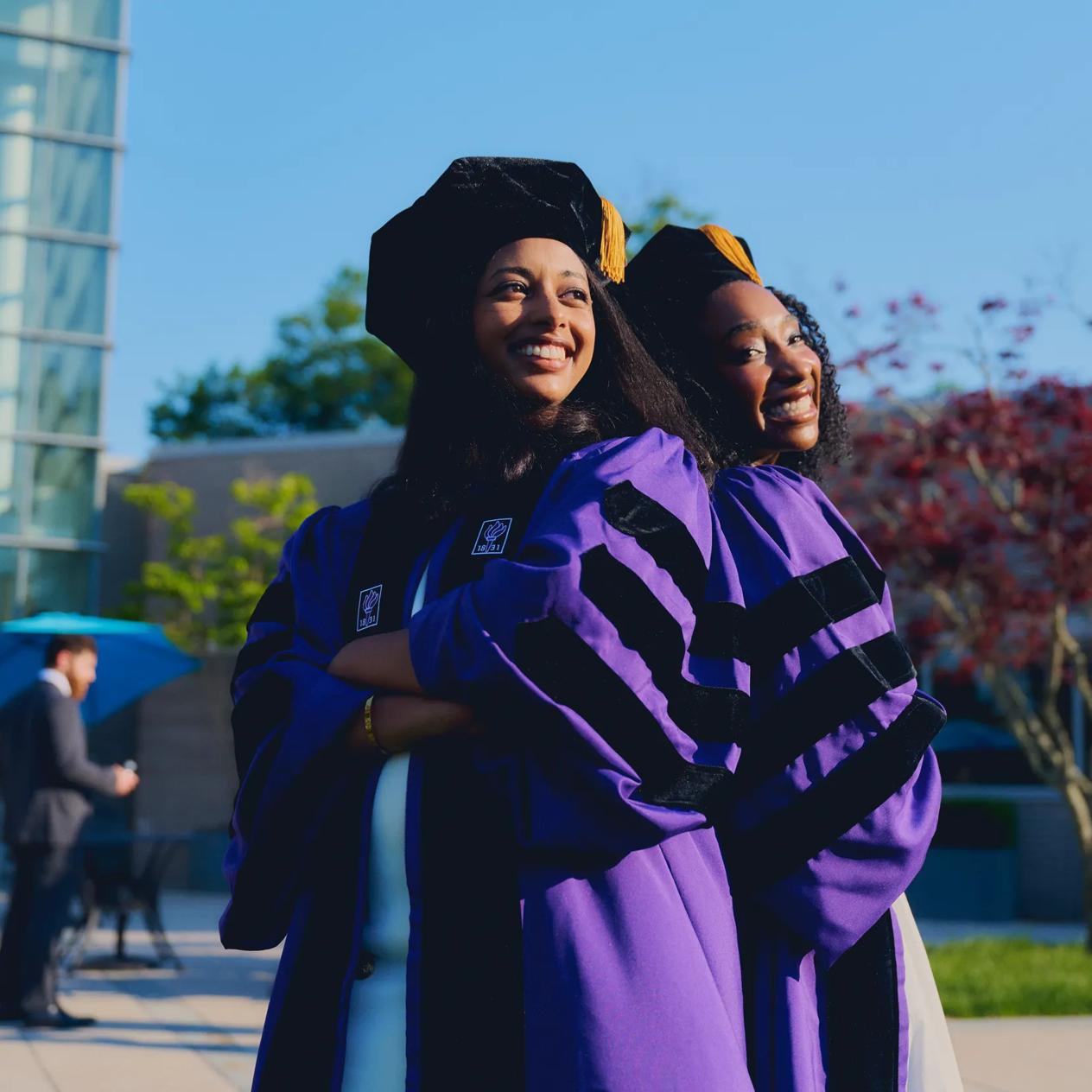 Two female NYU Grossman Long Island School of Medicine graduates stand outside on a sunny day with huge smiles. They are back-to-back, with cross arms, wearing purple commencement robes and doctoral hoods.