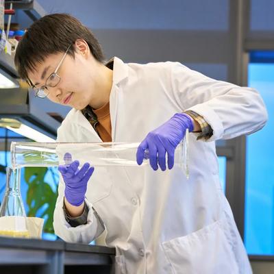 A researcher stands and pours a liquid into a container in a research lab.