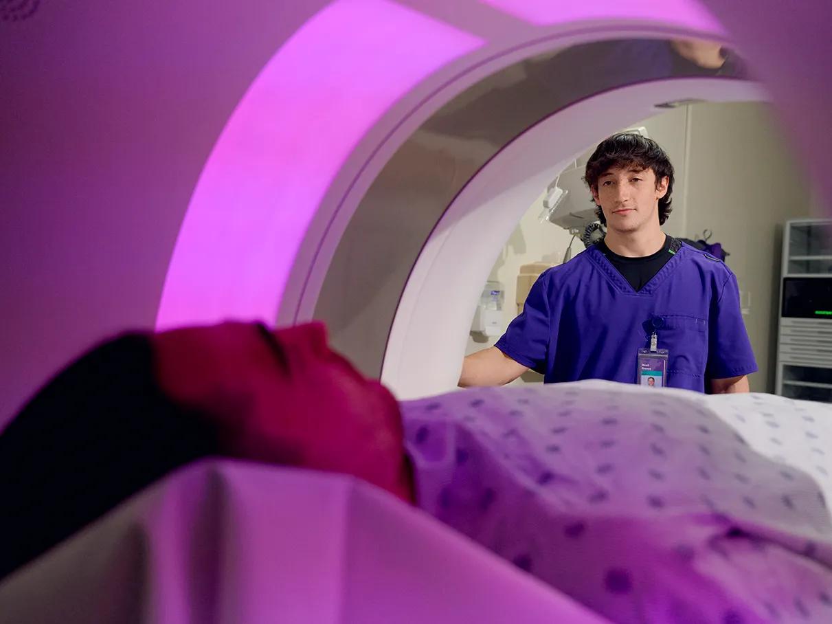 A male patient, forward left, lies down in an MRI machine. An MRI technologist, standing right, presses a button on the machine.
