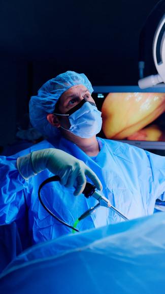 A male NYU Langone doctor wearing blue hospital scrubs, a mask, and a face shield, performs surgery on a patient.