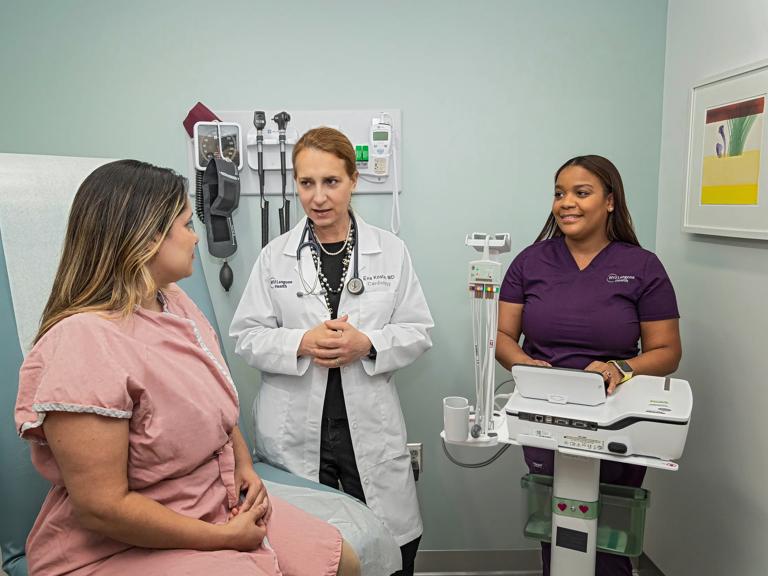 A female patient, left, sits on a patient bed in an exam room. A female doctor, middle, and a nurse, right, stand in the room next to her.