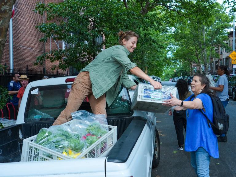 A woman, left, stands in a truck and hands a crate to another woman, right, standing on the street.