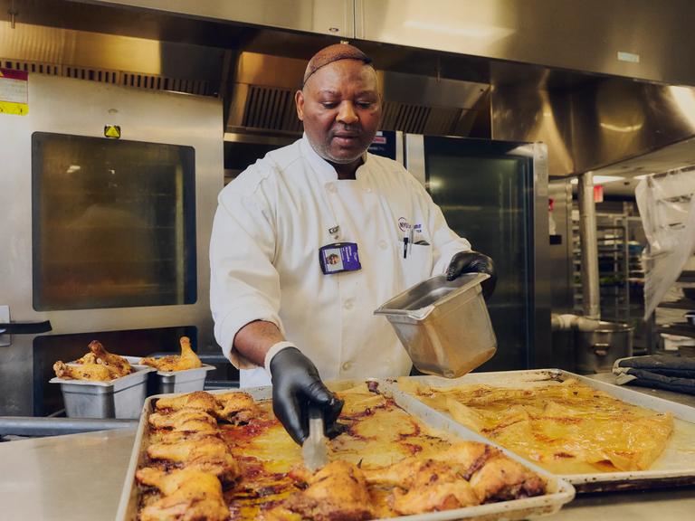 A cook stands in a kitchen over a tray of chicken.