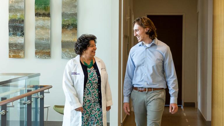 Dr. Gladys Ayala, Dean of the NYU Grossman Long Island School of Medicine, smiles while she walks and converses with a student wearing a white coat.