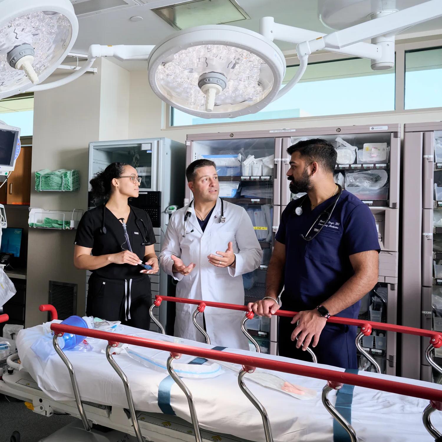 An NYU Grossman School of Medicine doctor, flanked by two nurses in dark blue scrubs, talk over a wheeled hospital bed with rails.
