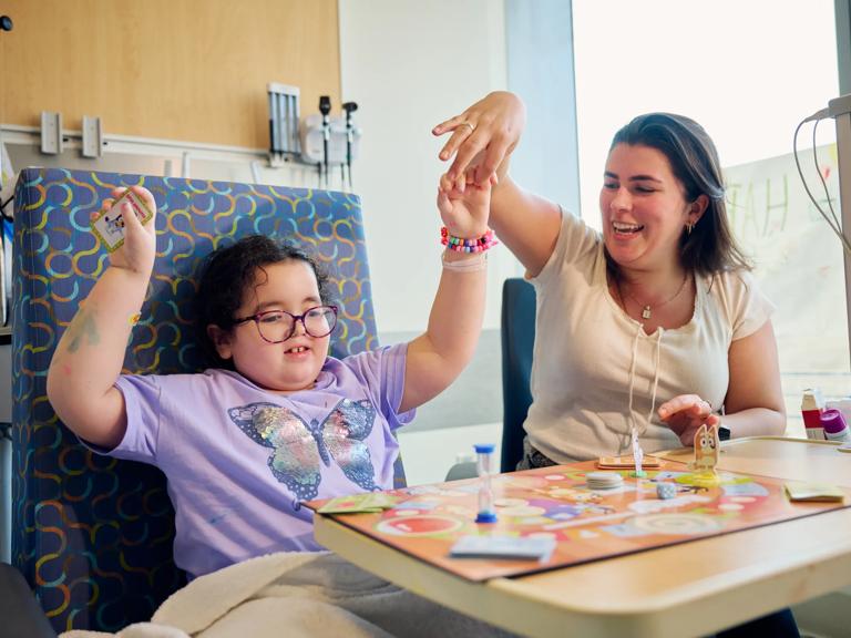 A child, left, sits in an inpatient room while raising her arms and holding a woman&rsquo;s hand.