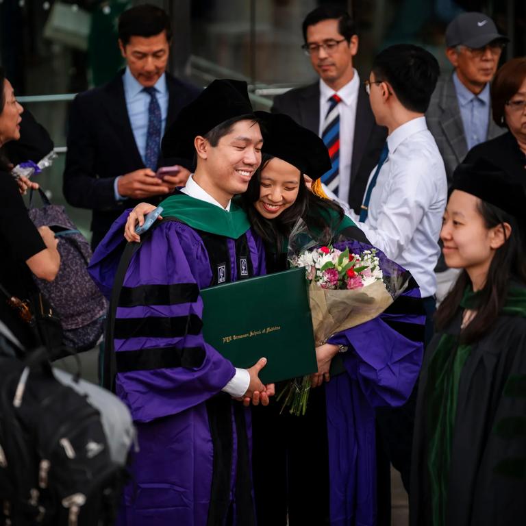 A male and female graduate wearing a purple commencement robe and doctoral hood share a smiling embrace outside Grossman School of Medicine. The man holds his degree, while the woman holds a bouqet of colorful flowers.
