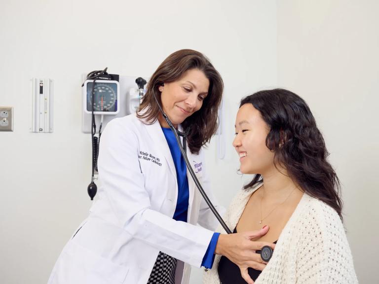 Michelle Bloom, MD, left, stands while using a stethoscope on a female atient, right, seated in an exam room.