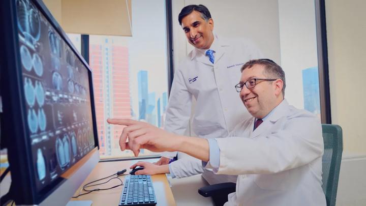 Rajwanth Veluswamy, MD, left, stands next to Paul E. Oberstein, MD, right, sitting and pointing at a computer monitor.