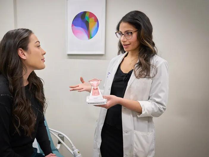 A female doctor, right, stands and holds an anatomical model while speaking to a patient, right, seated on an exam bed.