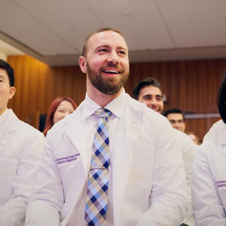 A Grossman Long Island School of MEdicine student stands with other students wearing white coats, smiling as they listen to a speaker.