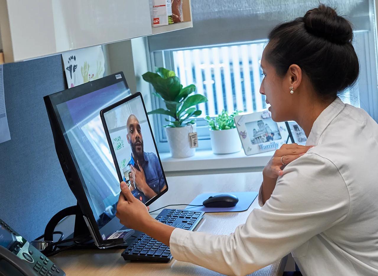 A doctor, right, sits at a desk in an office while holding a tablet and conducting a video call with a patient.