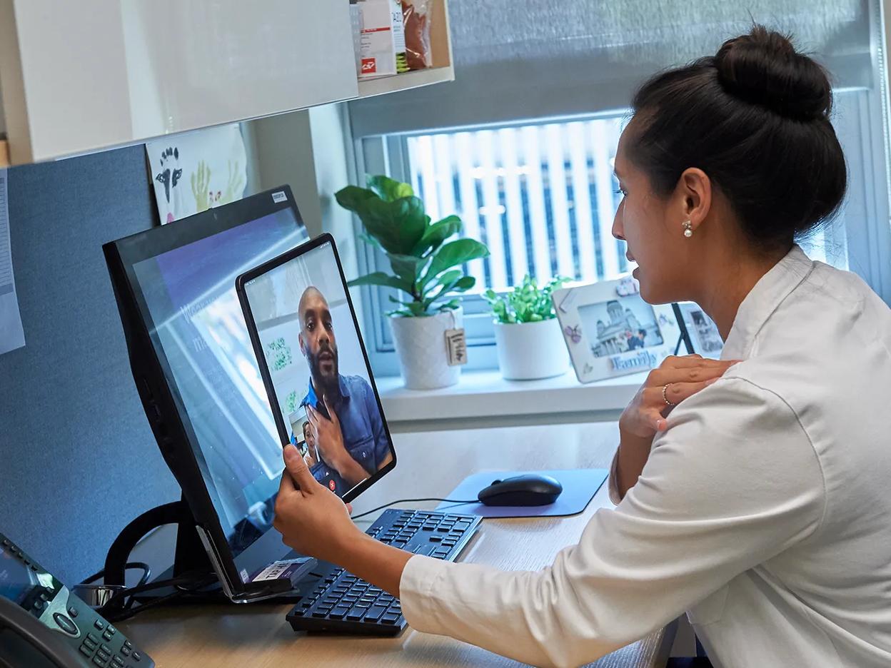 A doctor, right, sits at a desk in an office while holding a tablet and conducting a video call with a patient.